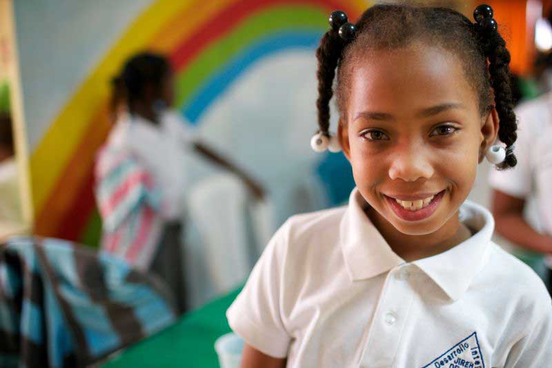 A girl smiles in front of a rainbow-painted wall in her classroom