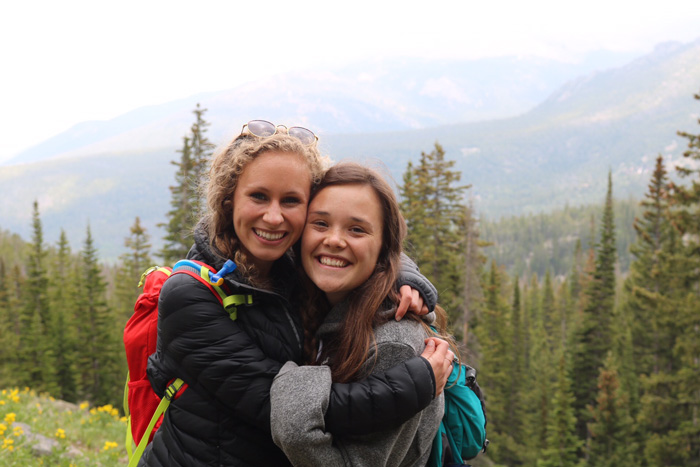 Two young ladies hug and smile with mountains behind them