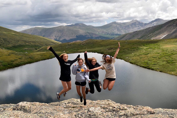 A group of young ladies jumping with mountains and a lake behind them
