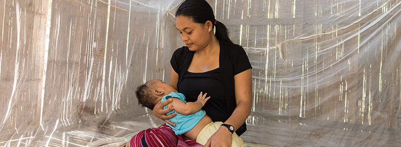 A mother holds a baby while sitting on a bed underneath a malaria bed net