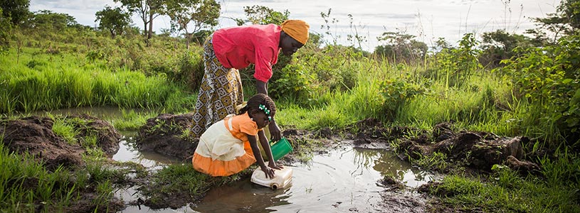 Two Ugandan women fill buckets with water from a pool of dirty standing water.