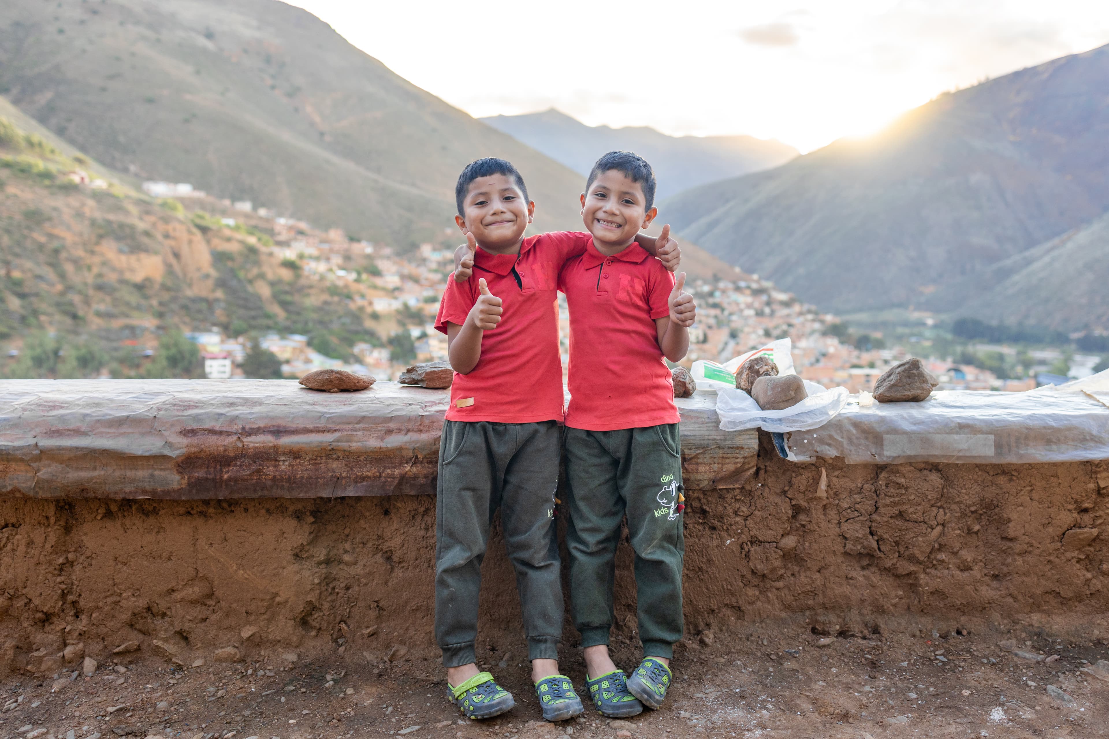 Two twin boys wearing red polos stand in front of rolling hills and a sunset while giving a thumbs up.