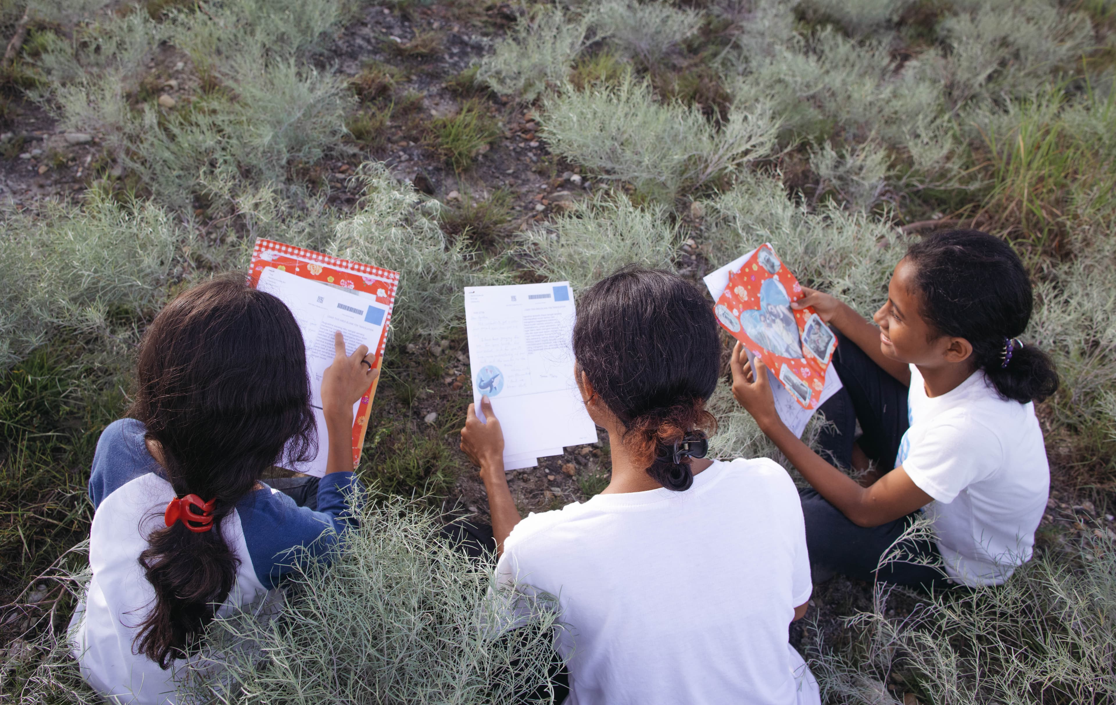 Three Indonesian girls in white T-shirts sit in a field reading letters.