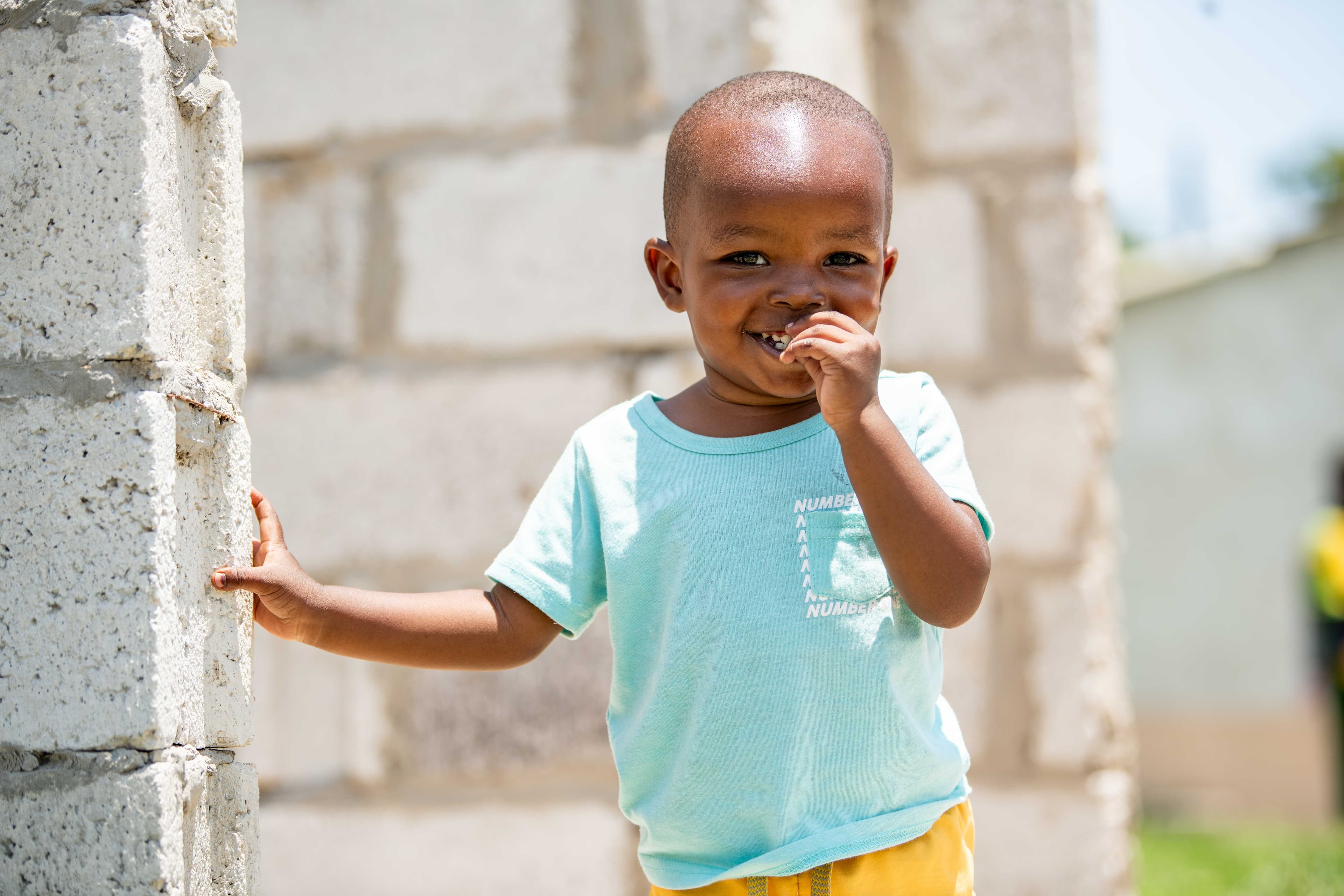 A young boy wearing a bright blue shirt smiles for the camera with his hand in his mouth.