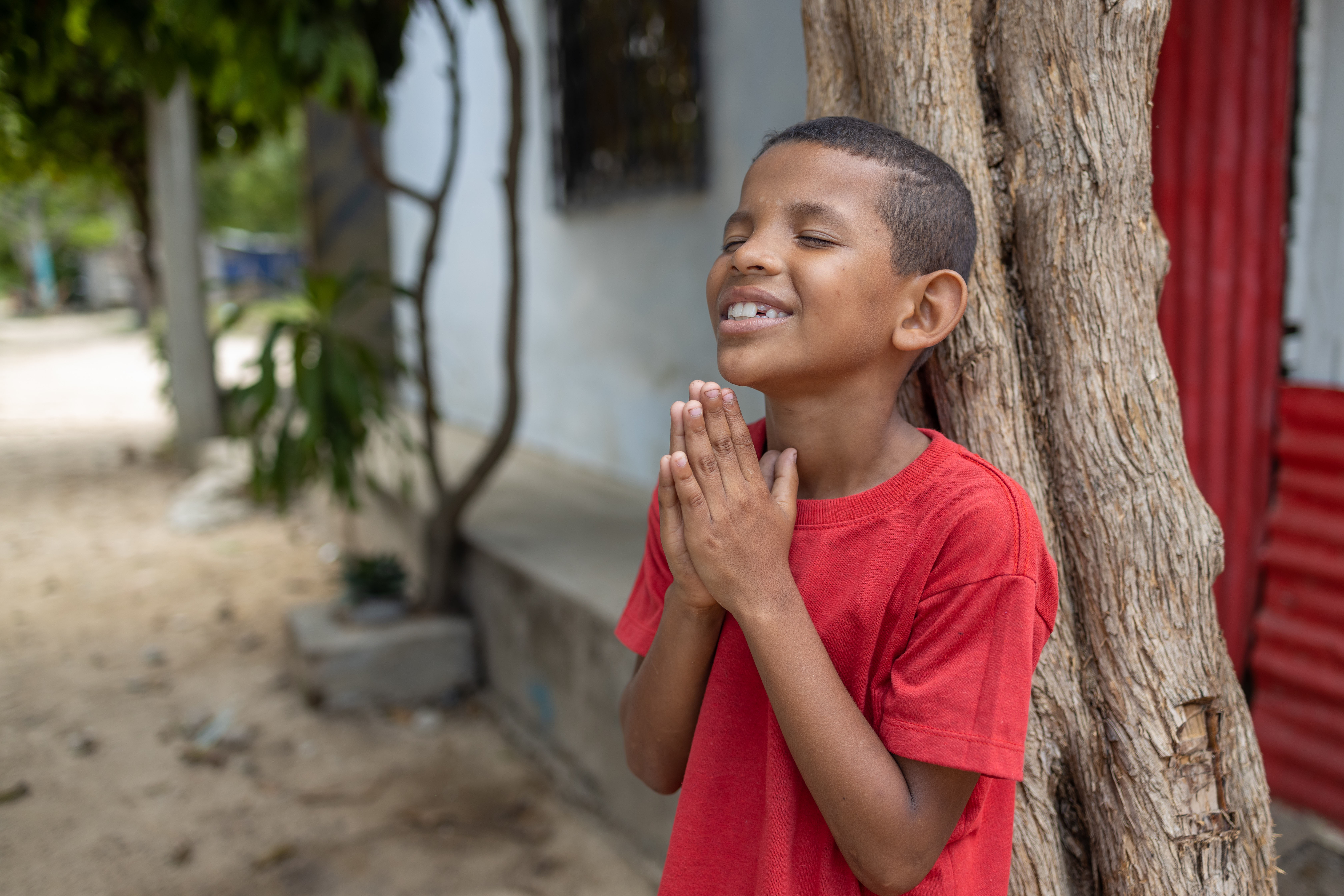 Young boy wearing a red shirt leans on a tree with his hands clasped in front of him as he prays and smiles.