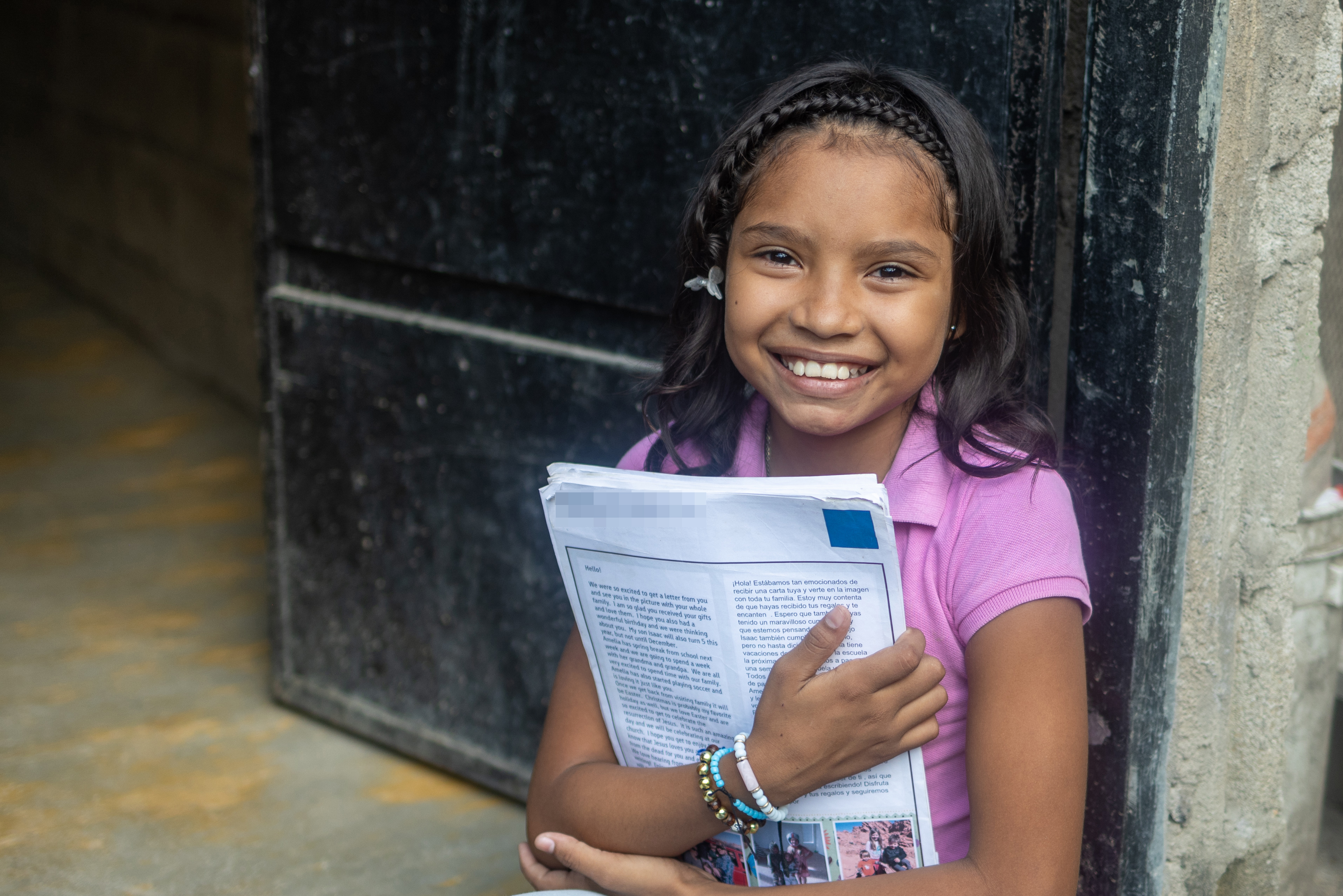 A young girl wearing a pink shirt holds a letter and smiles for the camera.