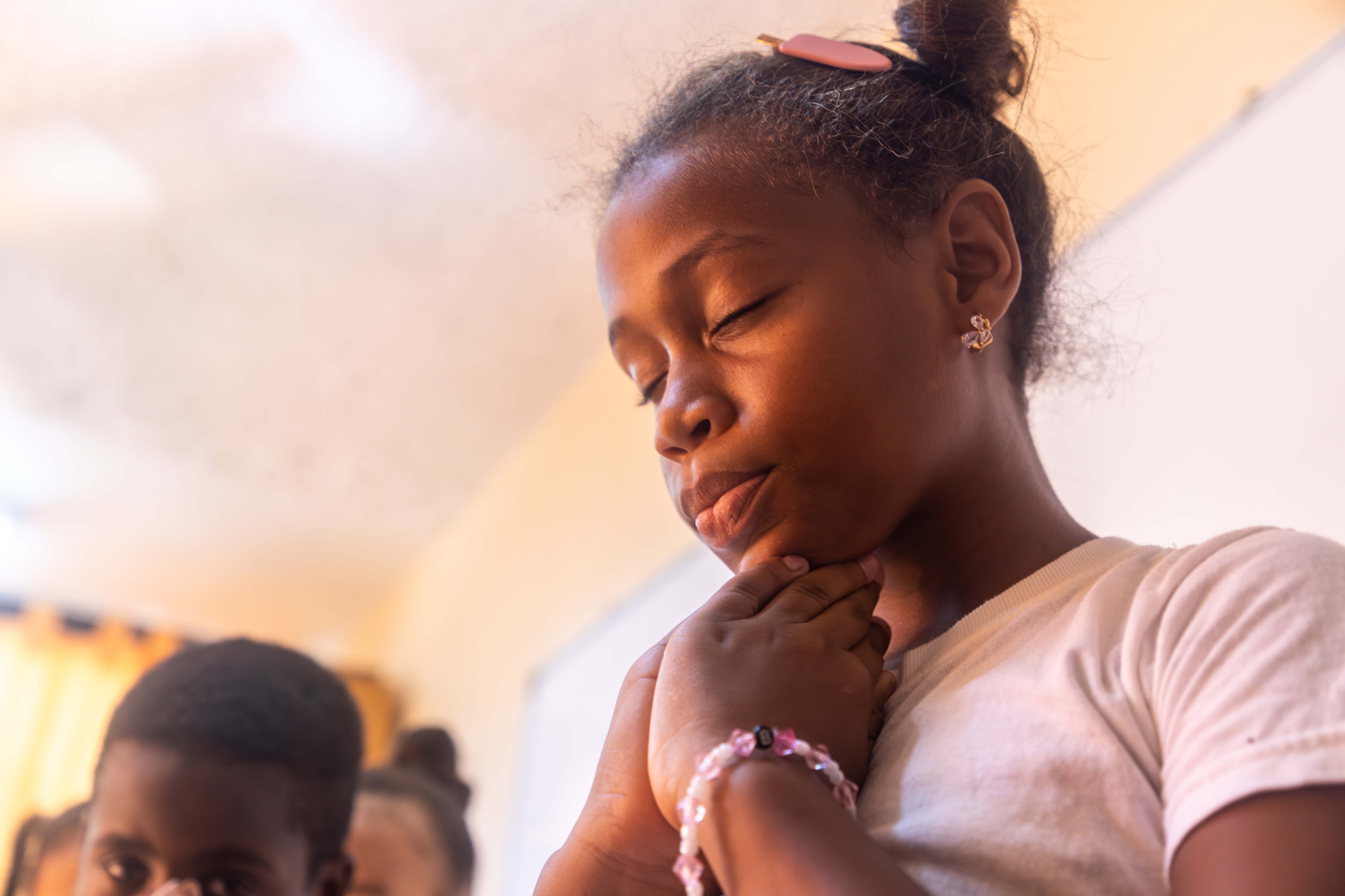 Young girl wearing a pink shirt closes her eyes and bows her head with her hands clasped in front of her.