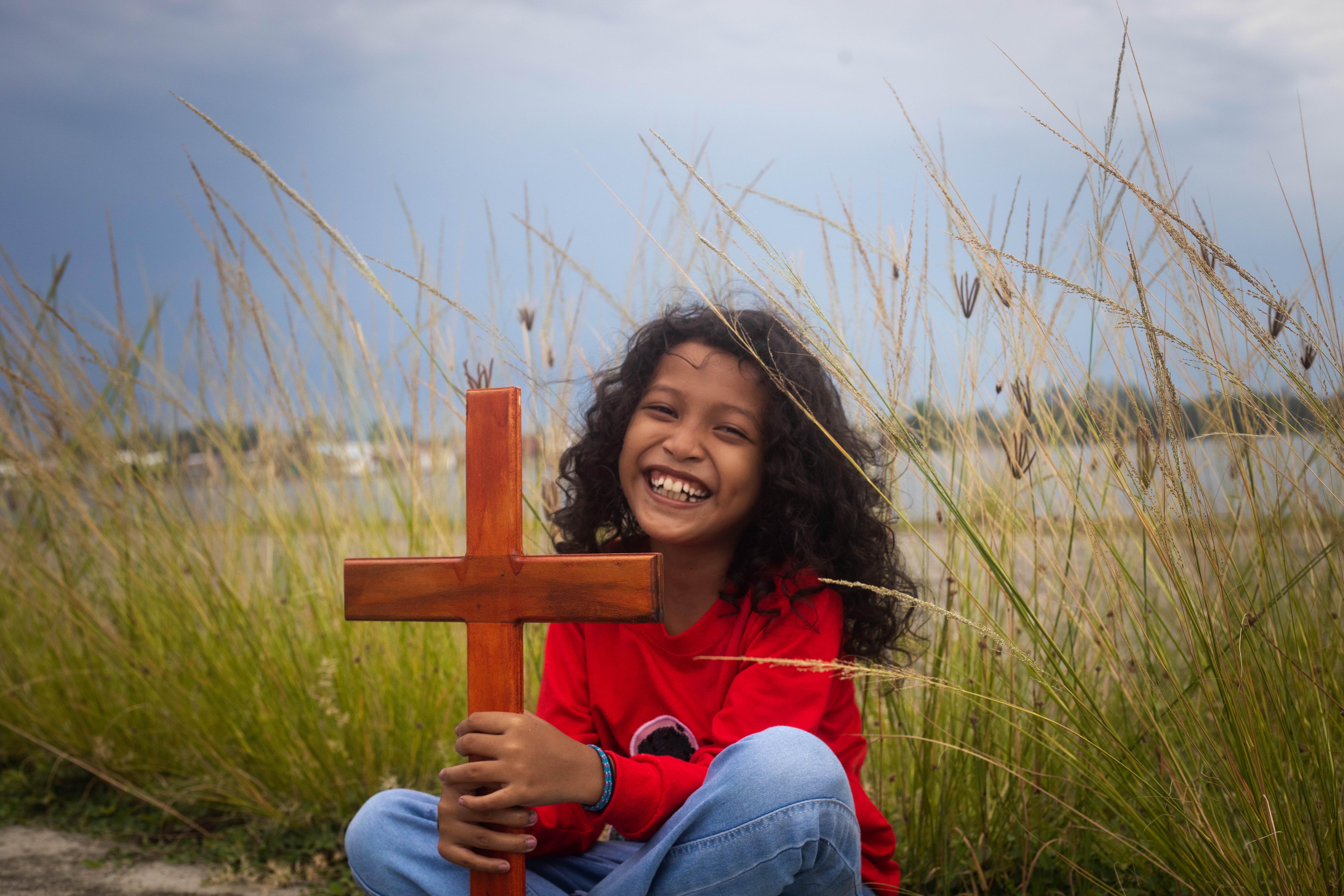 Young girl in a bright red shirt sits in front of tall grass holding a wooden cross and smiling for the camera.