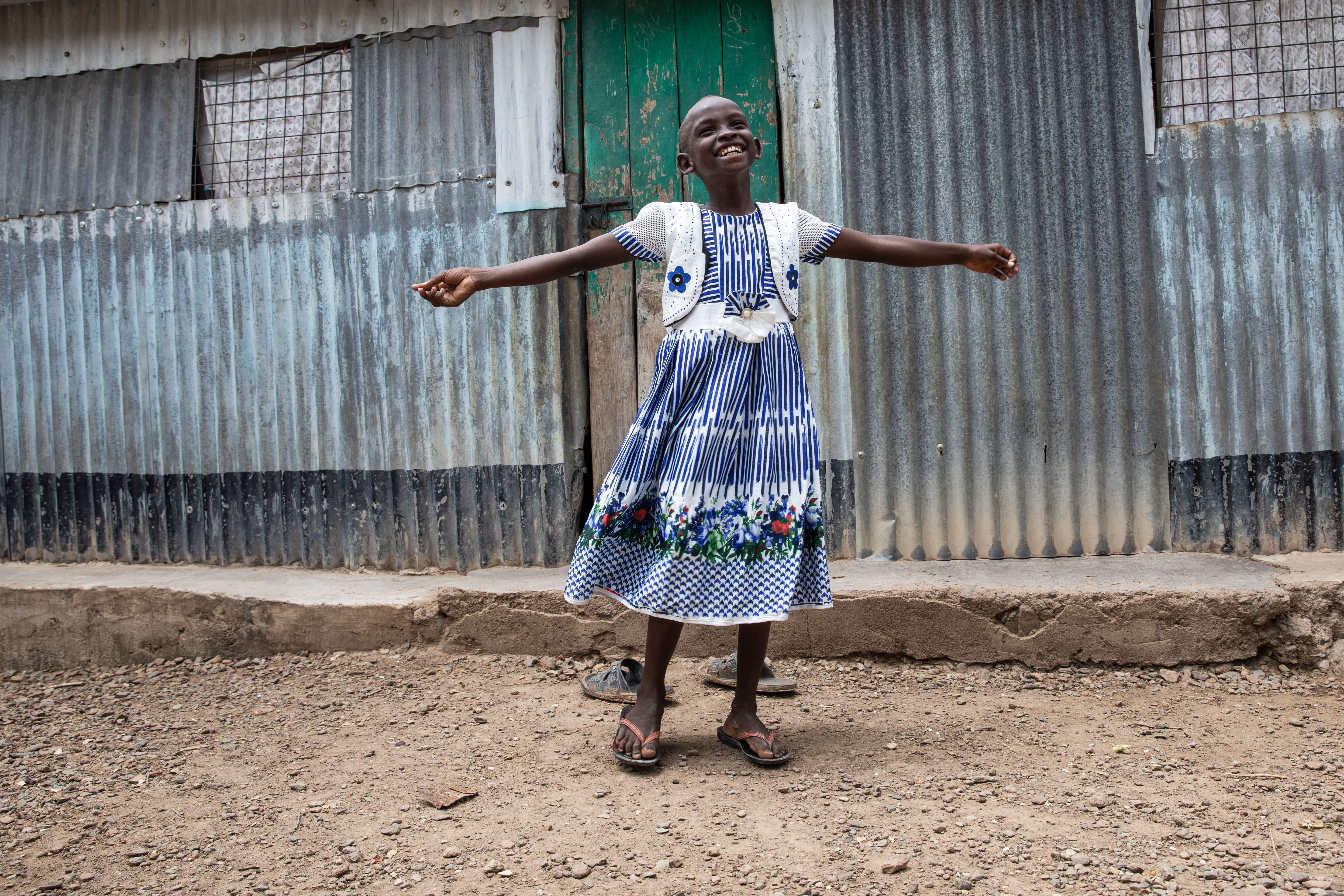  Young girl stands on a dirt road wearing a blue and white striped dress while smiling with her arms in the air.