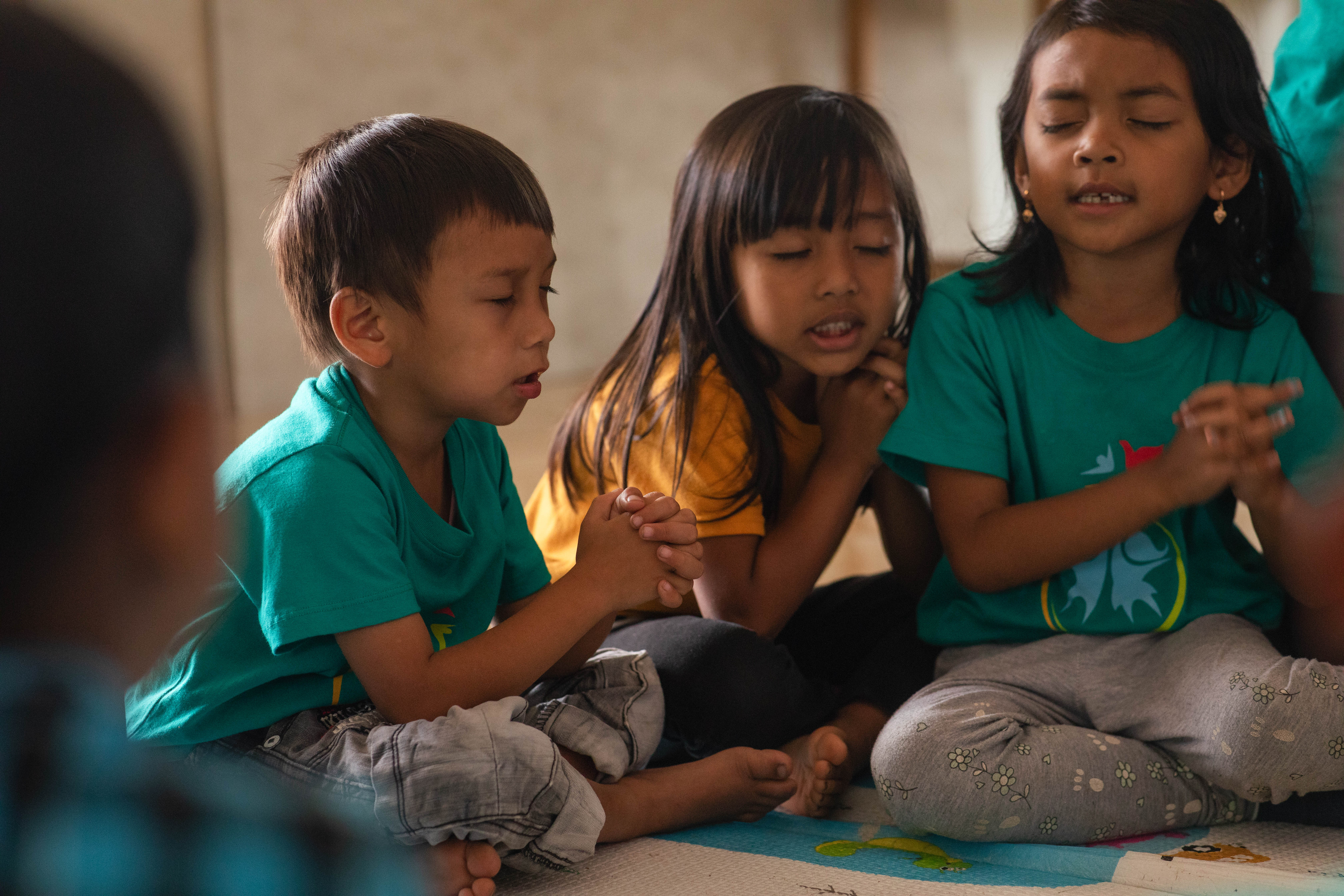 Three children sit on the floor with their hands clasped together and eyes closed as they pray.