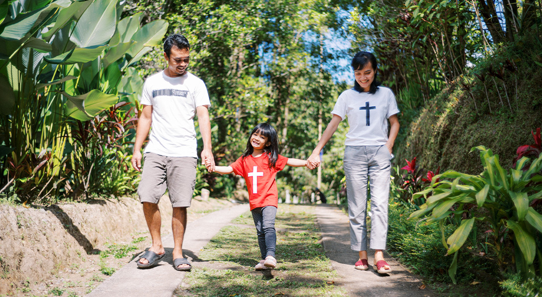 A mom and dad hold hands with their daughter while walking down a path.