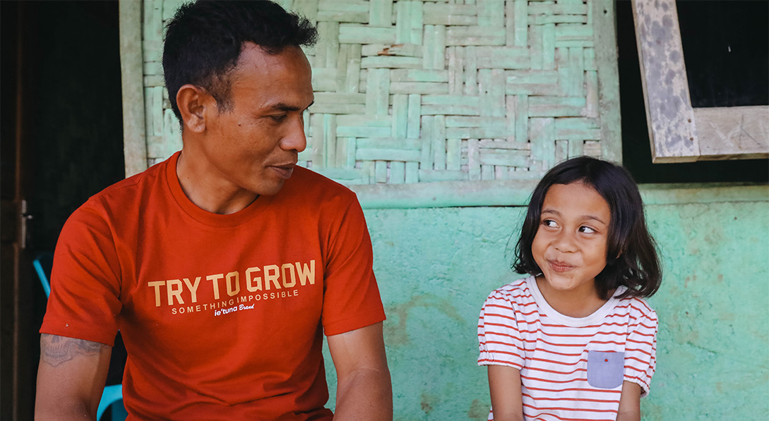 A dad in a bright red shirt sits next to his young daughter and talks to her. The daughter smirks at her dad.