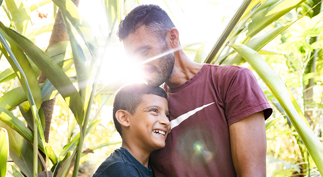 A dad hugs his son as the son smiles. Lush green plants surround them and a bright light shines behind them.