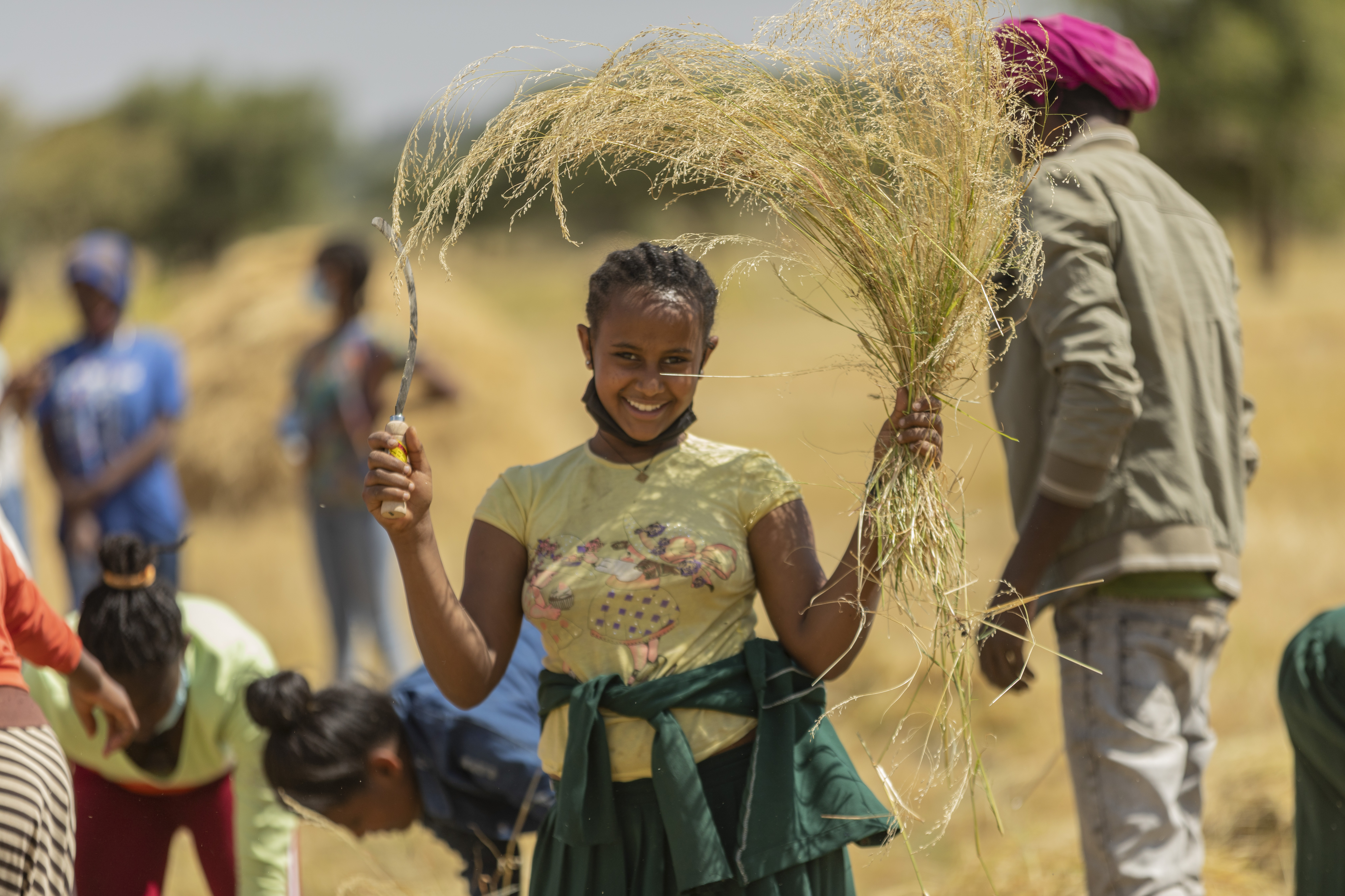 Young girl holds a sickle in one hand and a plant she harvested in the other while smiling for the camera.