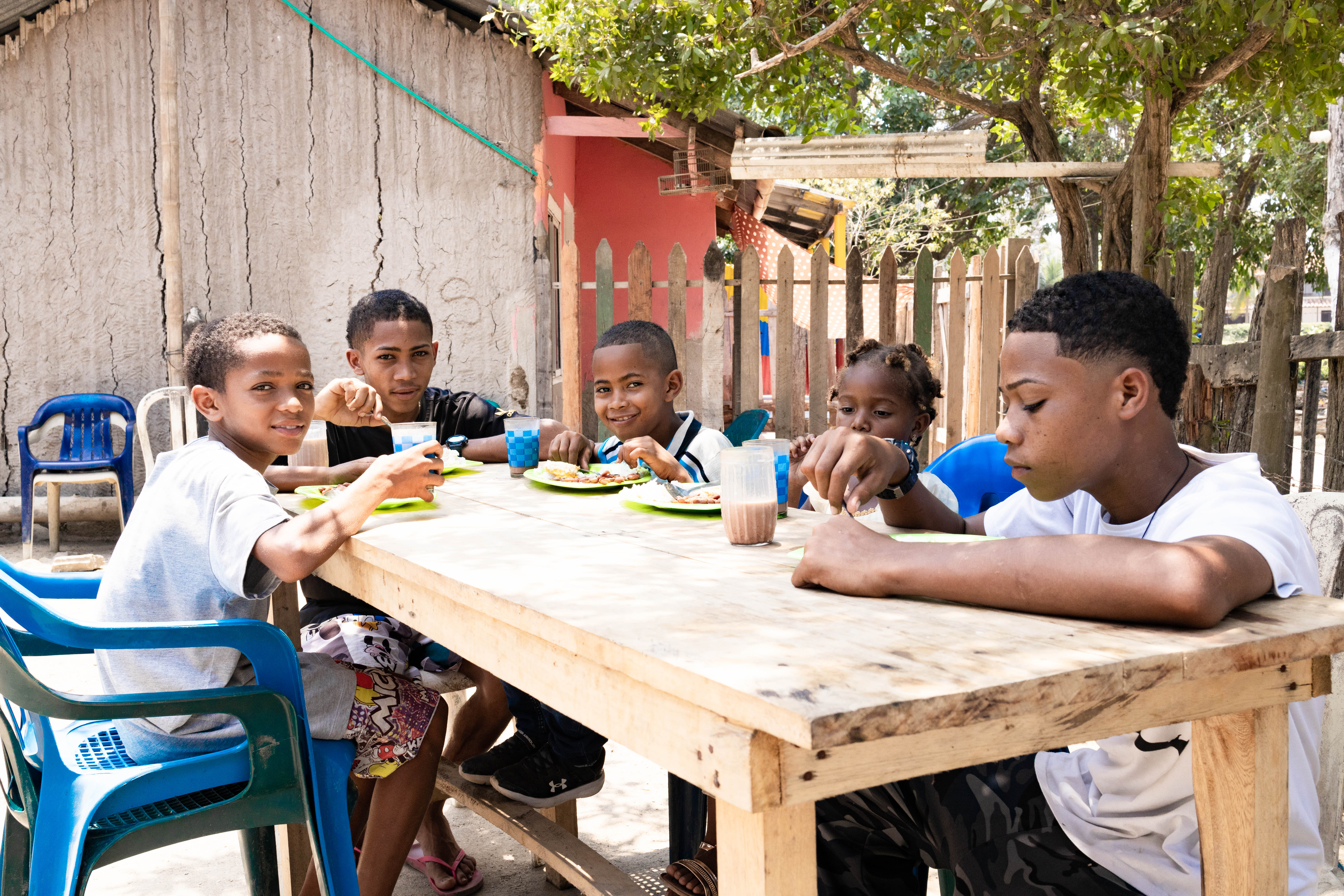 Five children sit at a wooden table and eat from green plates.