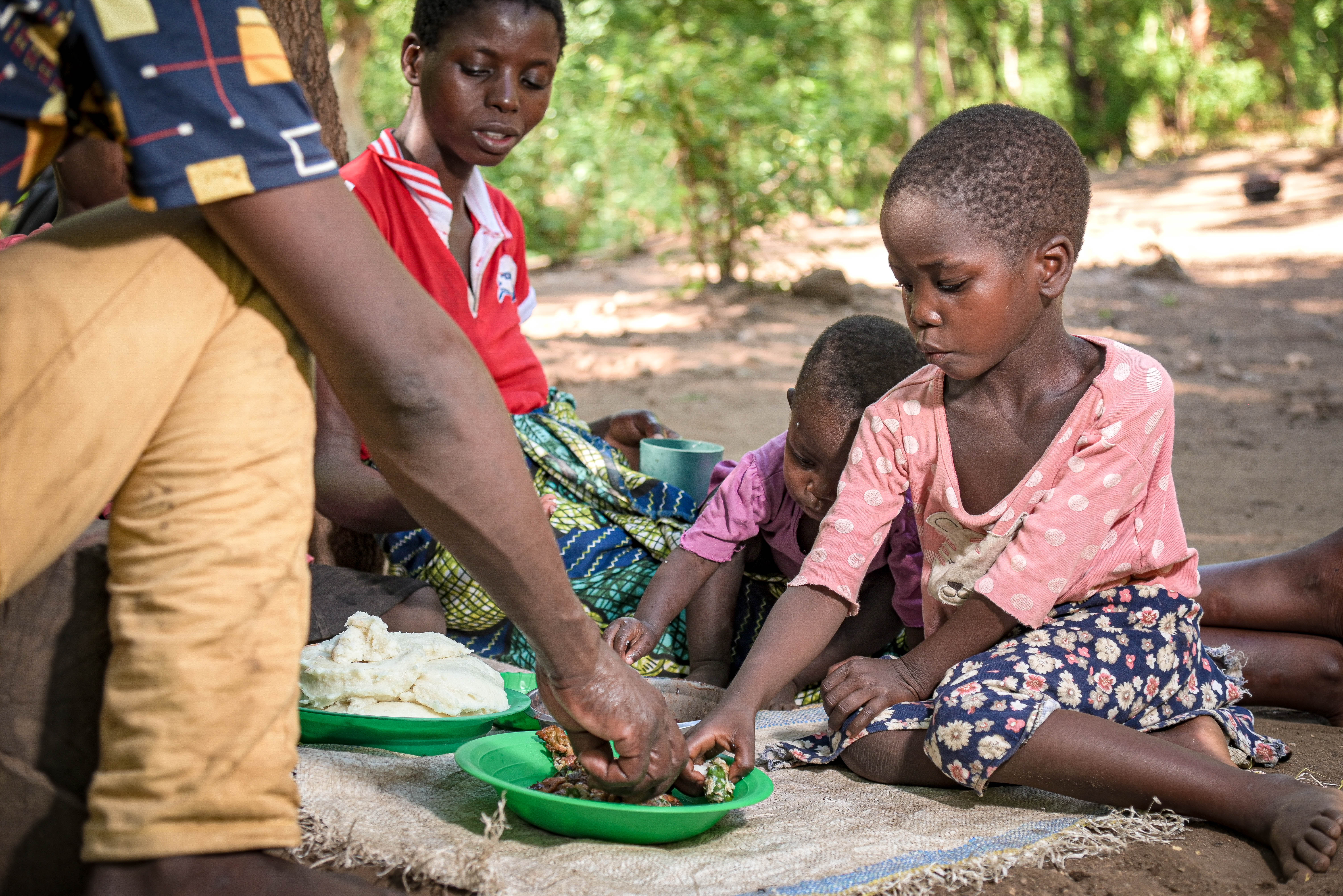 A young girl sits on a mat while eating from a green bowl.