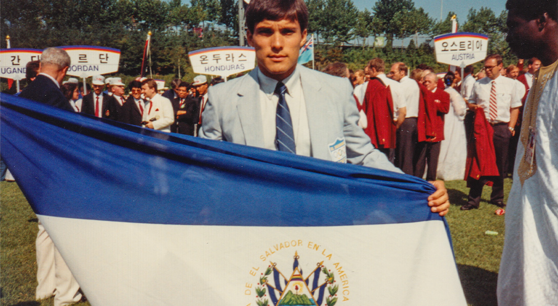 Jimmy Mellado stands behind the flag of El Salvador in a blue suit.