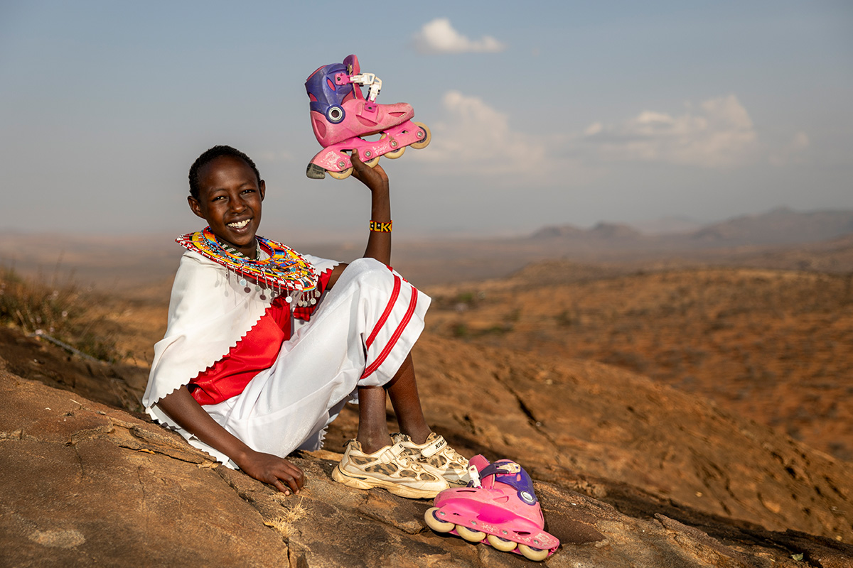 Young girl named Abigail smiles and sits on stony ground proudly lifting her bright pink and purple roller skate in the air.