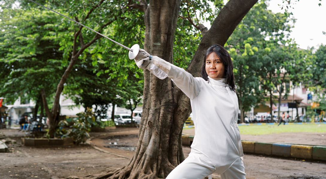 Elsya stands proudly wearing her white fencing gear and pointing her fencing sword into the air.