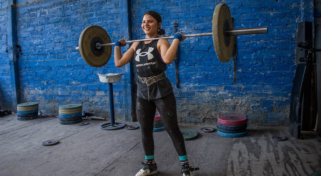 While smiling, Nicole stands on the wooden gym floor and lifts a barbell into the air.
