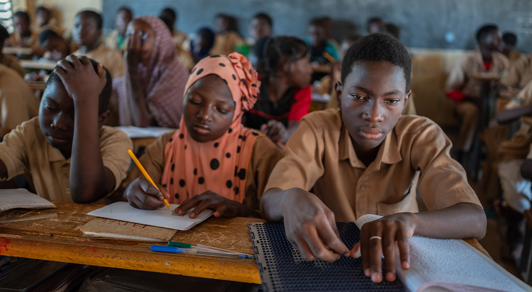 a boy sits by other students at a desk with and uses hands to read braille