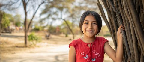 Young girl sits in a field and looks to the right while smiling widely.