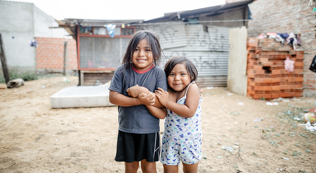 Two young girls stand together, arms linked, smiling for the camera in front of their home.