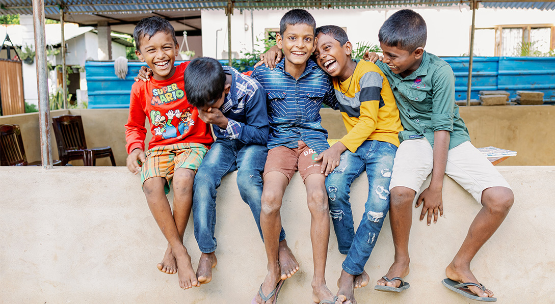 A group of five young boys sit on a wall while laughing and smiling for the camera.