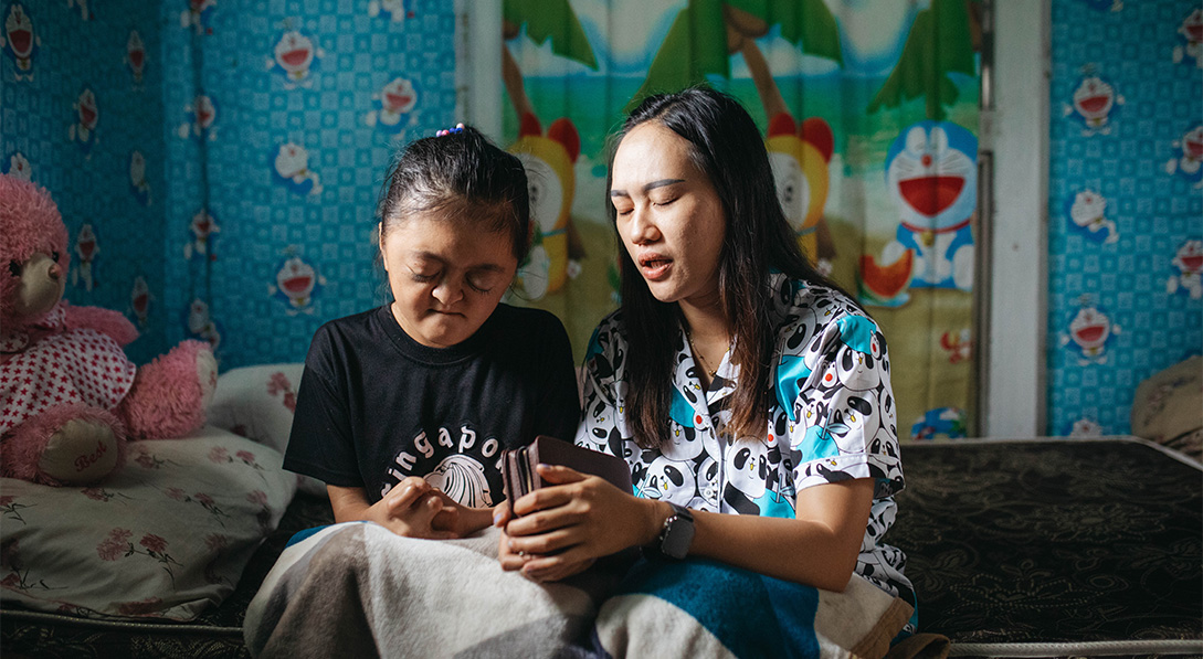 A young woman and a young girl sit on a bed with their eyes closed as the young woman prays while holding a Bible.