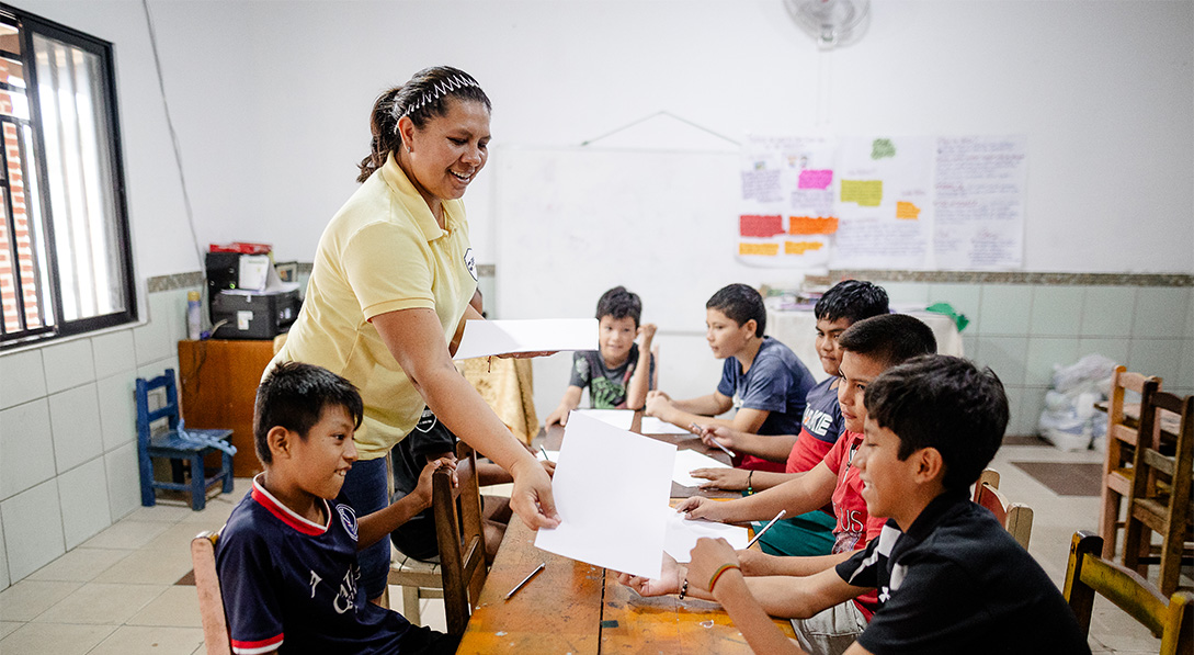 A young woman wearing a yellow shirt passes paper to young boys sitting around a wooden table.