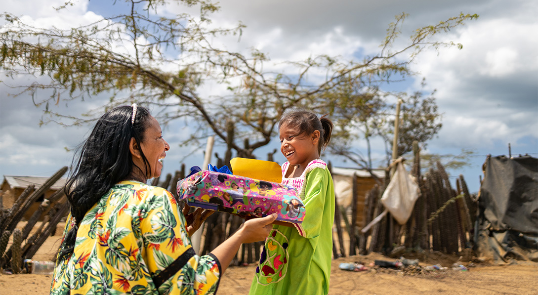 A woman wearing a brightly colored shirt hands a wrapped gift for a young girl wearing a bright green dress. Both of them smile brightly.