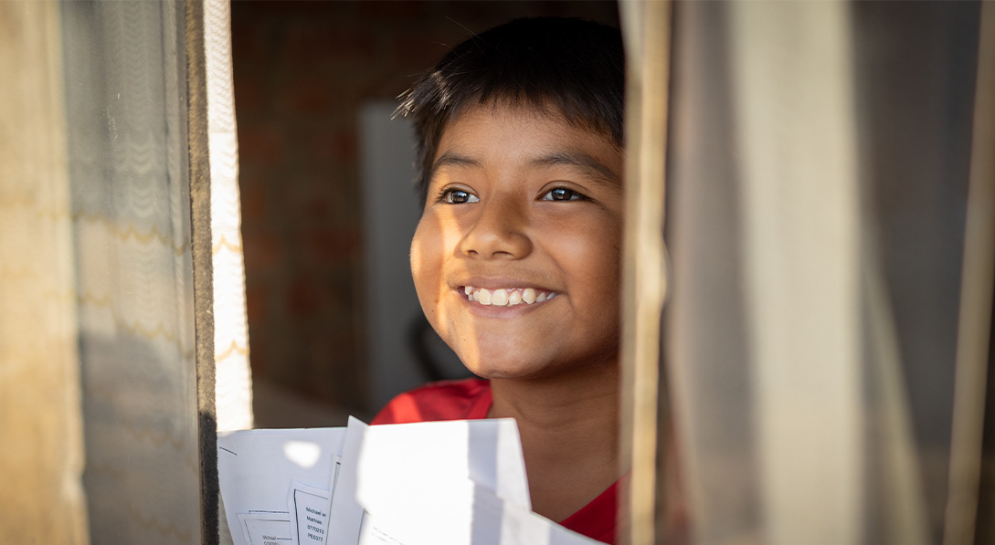 A young boy looks out of a window while holding a letter from his sponsor and smiling brightly.