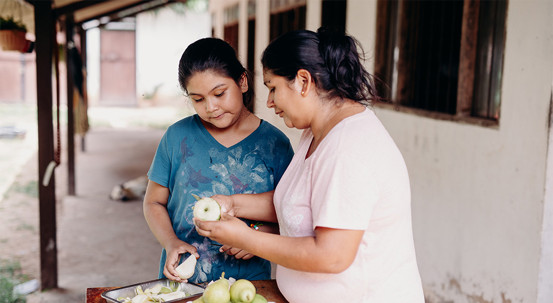 A woman and young girl stand side by side cutting and slicing green apples outside of their home.