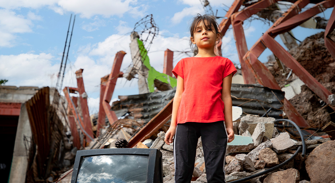 A young girl wearing a bright red shirt looks at the camera while standing in front of a pile of rubble.