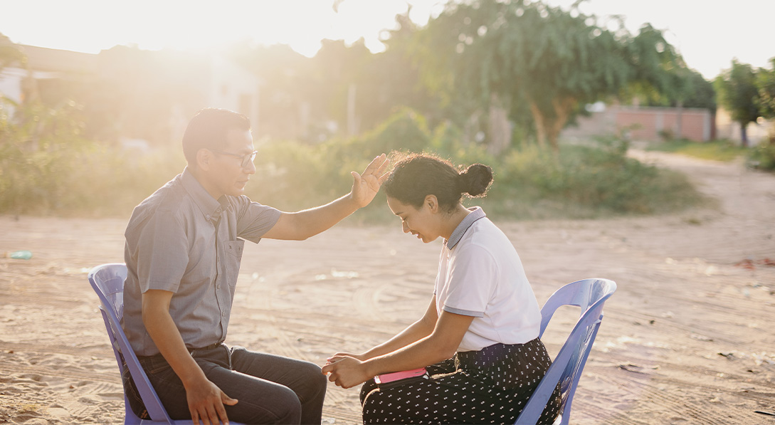  A pastor and a young woman sit across from each other. The pastor’s hand is raised above her head as they pray together. Light streams in from the left.