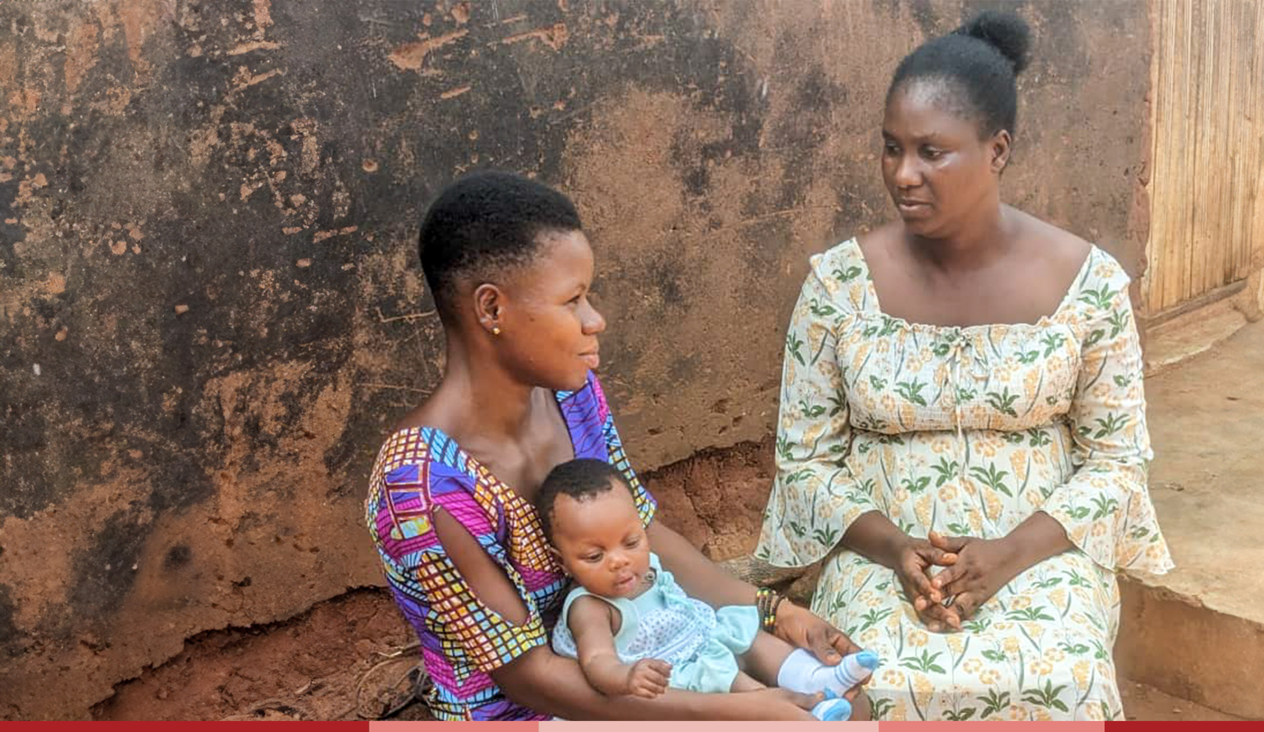 a mother and baby sit on a bench beside a young woman in a floral dress