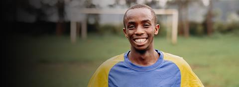 Teenager wears blue and yellow Rwanda soccer jersey and smiles at the camera.