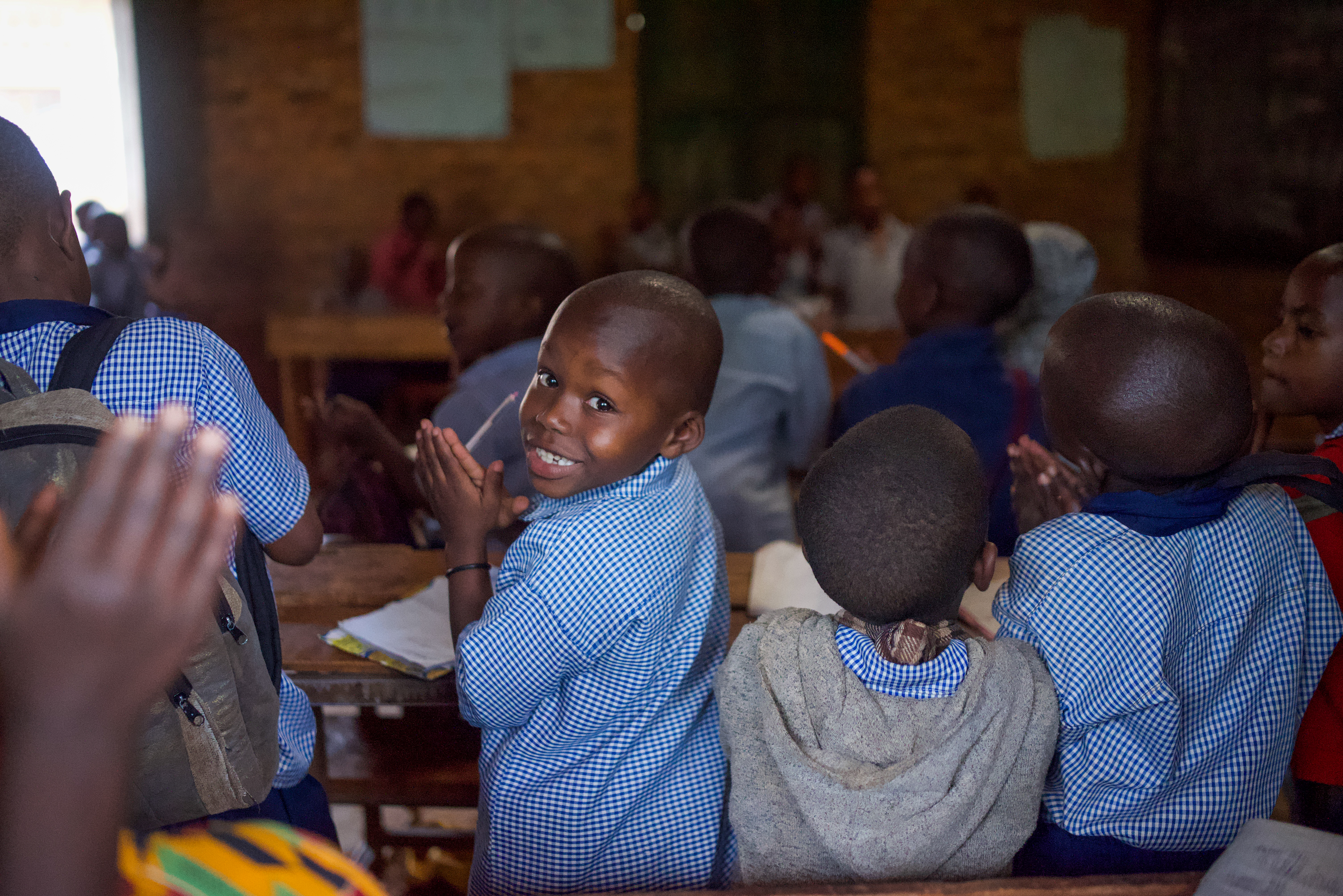 A young boy wearing a blue and white plaid shirt tuns his head to smile for the camera while surrounded by other children.