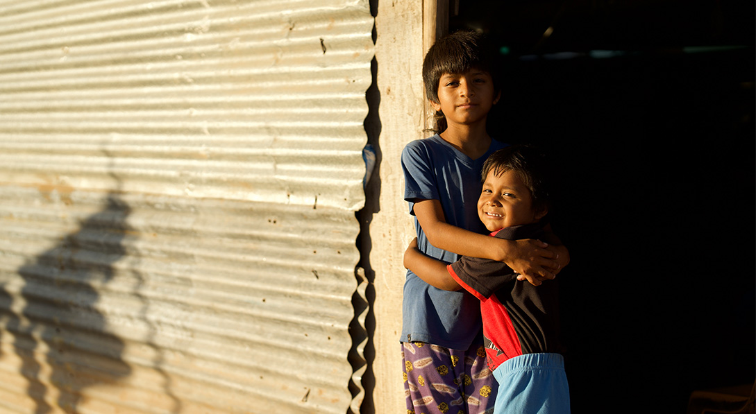 Two young boys stand in the doorway of their home hugging and smiling for the camera.