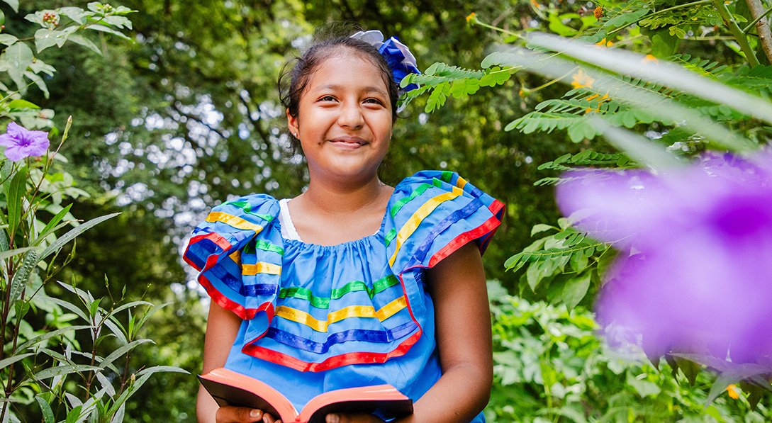 Young girl smiles at the camera while holding a Bible. She’s surrounded by flowers and lush green plants.