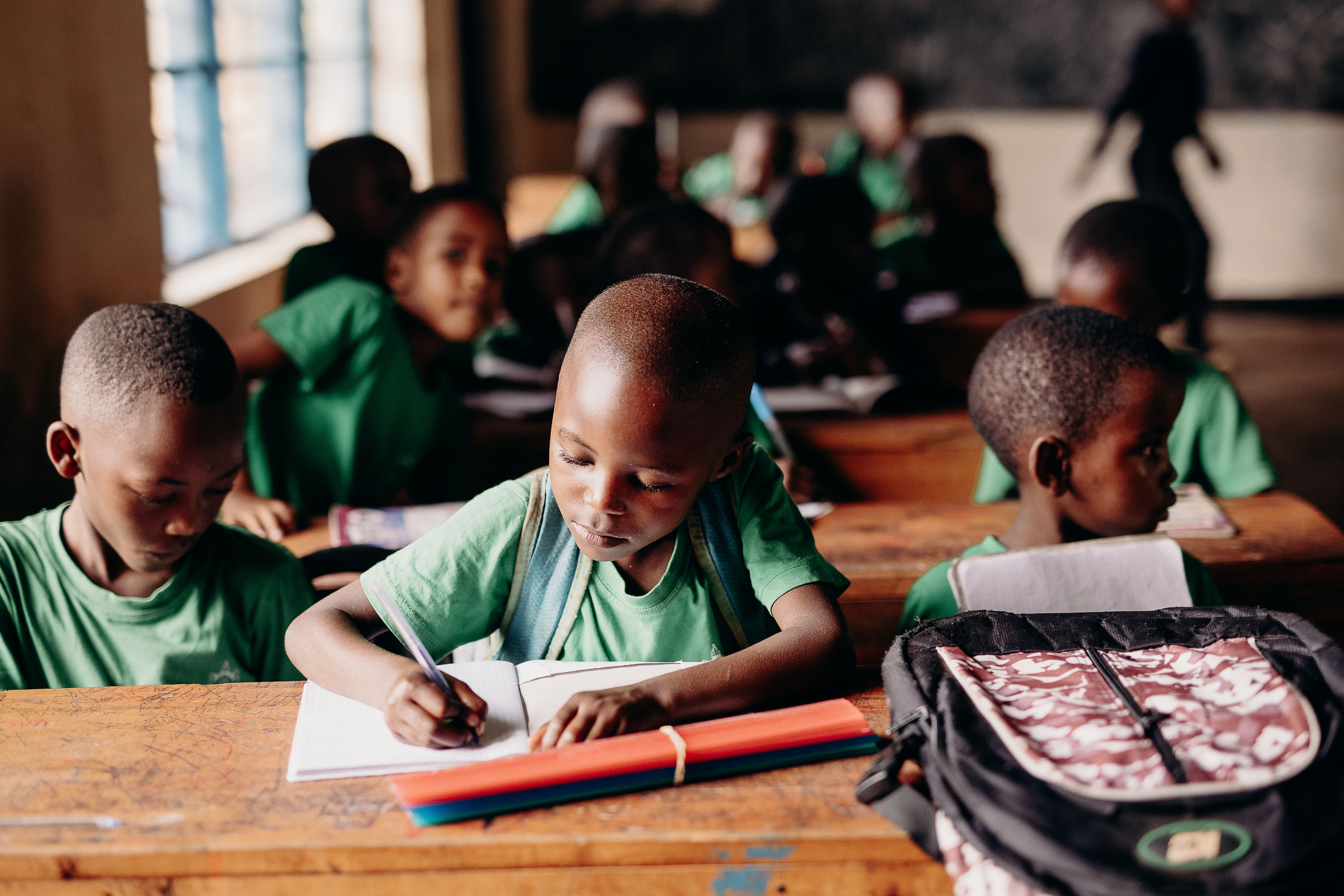 A young boy wearing a green shirt and a backpack sits at a desk writing in a notebook.