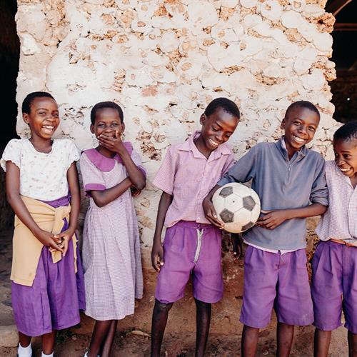 Five children wearing school uniforms stand in front of a stone building smiling and laughing. One of them holds a soccer ball.