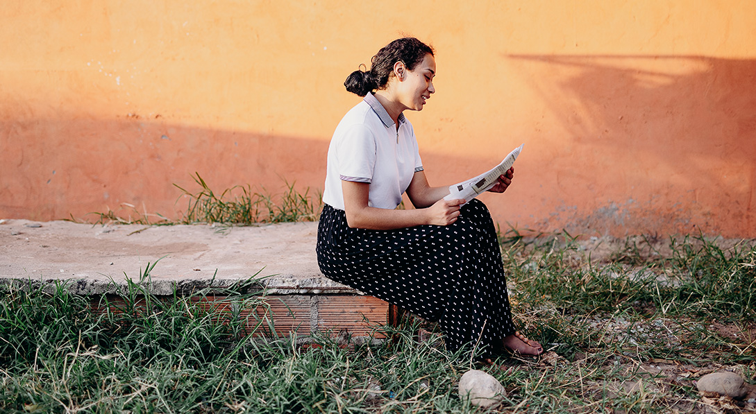 A young woman reads a letter while she sits outside a bright orange building. She is wearing a white blouse and black skirt and smiling.