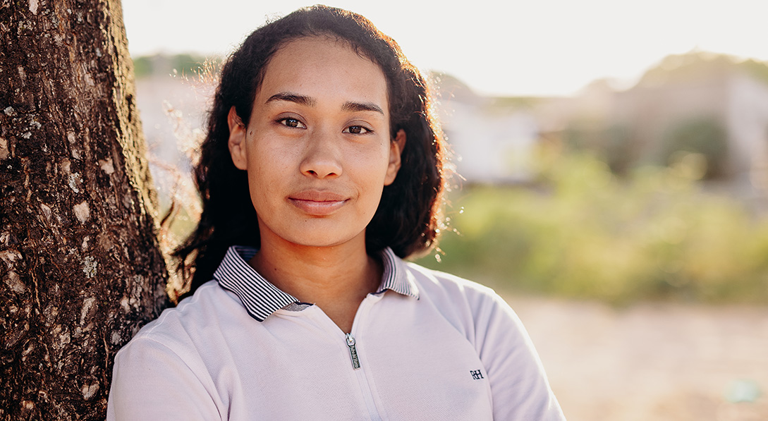 A young woman wearing a white blouse stands next to a tree and looks calmly at the camera.