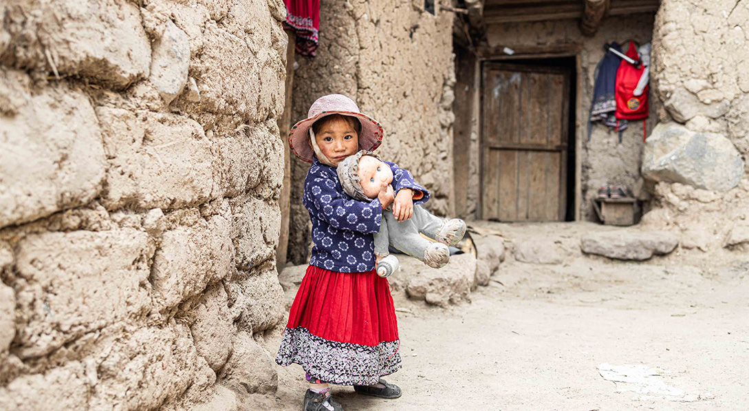 Little girl wearing a hat and a brightly colored dress stands outside her clay home holding a doll.