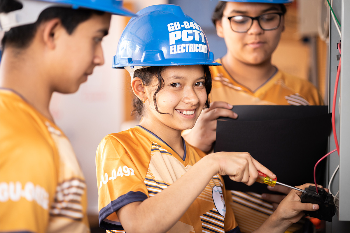 A young girl wearing a blue hard hat works on an electrical breaker and smiles at the camera.