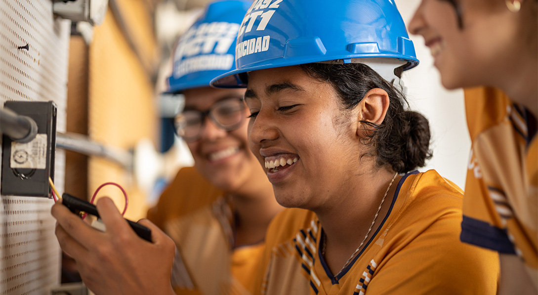 Three girls wearing blue hard hats smile and laugh as they work on an electrical circuit.