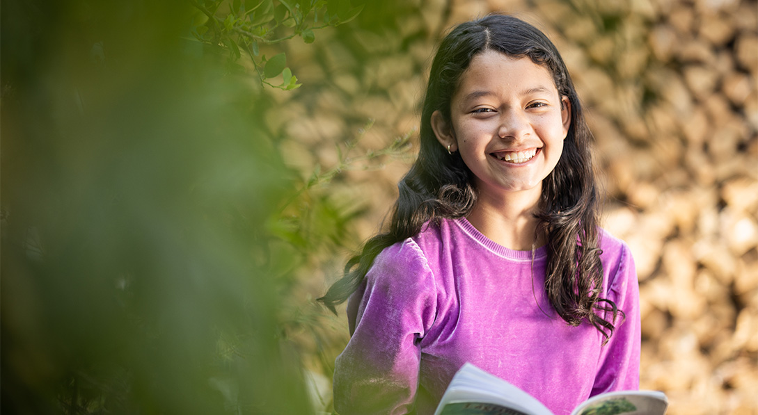 A young girl wearing a purple shirt and holding an open book smiles for the camera.