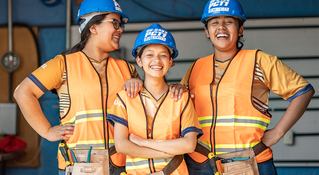 Three young girls wearing blue hard hats and safety vests smile for the camera.