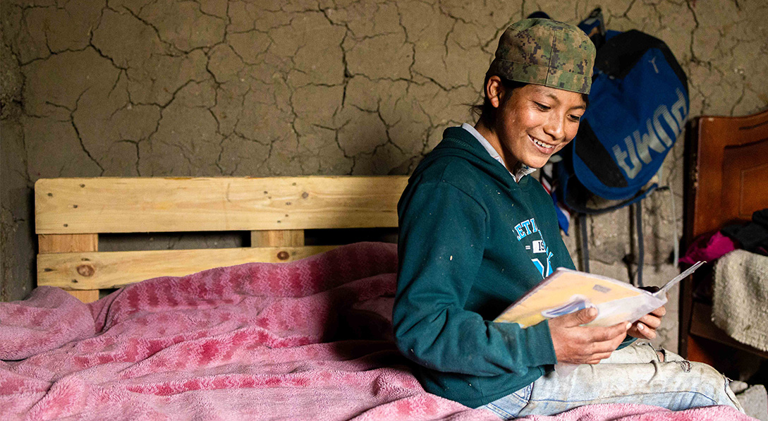 Young boy wearing a backwards camouflage hat sits on his bed and reads and book while smiling.